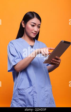 Ein gutaussehender Mitarbeiter des Klinikpersonals, der ein digitales Tablet nutzt, während er auf orangefarbenem Hintergrund steht. Schöne asiatische Krankenschwester Arbeiter mit modernen Tablette in der Hand. Studioaufnahmen. Stockfoto