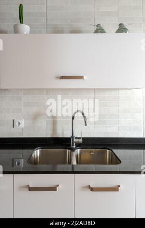 Modernes Küchenwaschbecken mit Doppelschale und Chromarmatur. Arbeitsfläche aus schwarzem Marmor und weiße Schränke. Minimalistisches Innendesign mit natürlichem Licht. Stockfoto