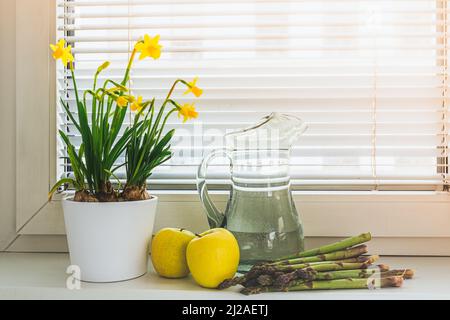 Frühlingsblumen Narzissen in Blumentopf, frische grüne Äpfel und Spargel auf Fensterbank. Konzept von Bioprodukten, gesunde Ernährung, Gartenarbeit Stockfoto
