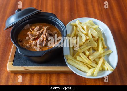 Geschmorte Eingeweide, Nieren, Niere und Fleisch in Soße, begleitet von gebratenen Kartoffeln auf dem Tablett in den Teller auf dem Holztisch. Stockfoto