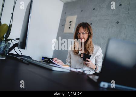 Eine kaukasische Frau mittleren Alters im Büro des Unternehmens schaut auf die Kamera, hält eine schwarze Tasse Tee oder Kaffee in der linken Hand und hält einen Stift in der rechten Hand. Stockfoto