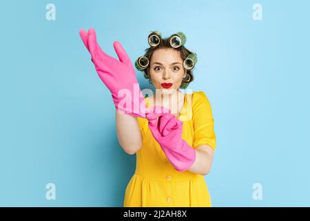 Schockiert junges hübsches Mädchen, Hausfrau mit Lockenwicklern auf ihrem Kopf isoliert auf blauem Studiohintergrund. Emotionen, Frauenrechte, Schönheit, Familie Stockfoto