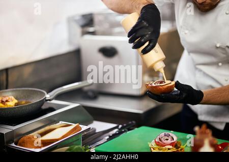 Blick auf die Ernte der Hände des Küchenchefs, der im Restaurant ein Brötchen hält und Sauce auf den Cheeseburger gießt. Männliche Hände in Handschuhen bereiten leckeren Rindfleisch Burger in der professionellen Küche. Konzept des Kochens von Lebensmitteln. Stockfoto