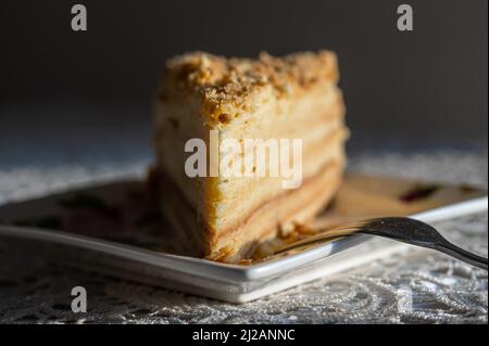 Nahaufnahme eines geschichteten Stücks des berühmten Napoleon-Kuchens, serviert auf einem quadratischen Teller mit einer Dessertgabel. Dreiecksscheibe mit knusprigen Krümeln illu verziert Stockfoto