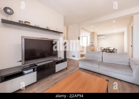 Abgewinkelte Ansicht der Wohnung mit Wohnzimmer, gepolstertem Sofa, Esstisch und offener Küche, Holzcouchtisch und großem Flachbildfernseher o Stockfoto