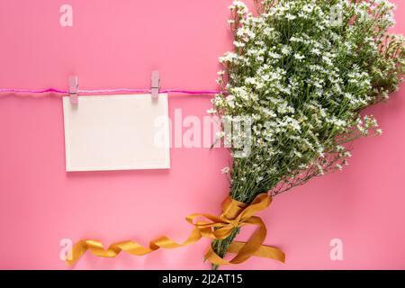 Mockup von pastellrosa Karte Notiz unbeschriftet Papier Blatt hängen an Seil mit Kleidung Stift und Frühling frischen weißen Blumenstrauß isoliert auf rosa Hintergrund. Stockfoto