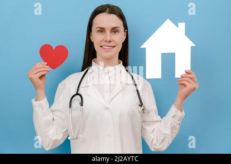 Glückliche junge Ärztin in professioneller medizinischer weißer Uniform und Stethoskop hält kleines Haus und rotes Herz, Blick auf die Kamera, isoliert auf blauen s Stockfoto