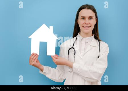 Junge Ärztin in professioneller medizinischer weißer Uniform und Stethoskop hält ein kleines Papierhaus, blickt lächelnd auf die Kamera und posiert isoliert über blu Stockfoto