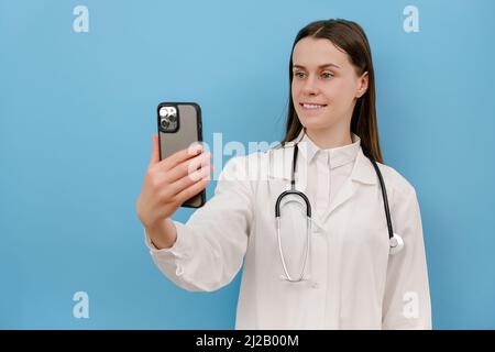 Lächelnde junge Ärztin hält Telefon Gespräch mit Patienten machen Telemedizin Online-Konferenz-Video-Anruf, isoliert auf blauer Wand. GP Remote consultatio Stockfoto
