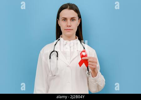 Die Ärztin hält ein rotes Band, schaut auf die Kamera, trägt Uniform und Stethoskop, isoliert an der blauen Wand im Studio. Bluttransfusion und Blutspende. Saum Stockfoto