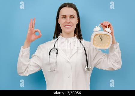 Porträt der schönen glücklichen jungen Frau Arzt hält Wecker, zeigt ok Zeichen, trägt weiße Uniform und Stethoskop, Blick auf die Kamera, posieren ist Stockfoto
