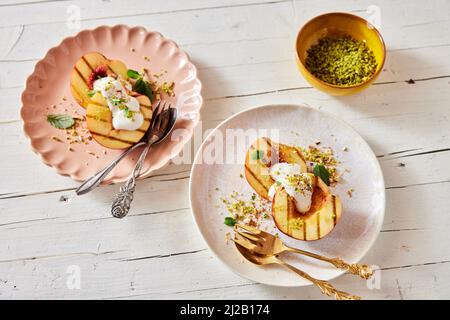 Draufsicht auf süß gegrillte Hälften von reifen Pfirsichen mit Joghurt und Minze, bestreut mit Pistazien, serviert auf Tellern auf einem Holztisch Stockfoto