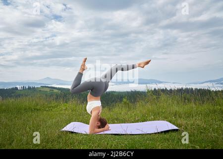 Aktive junge Dame trägt Sportkleidung in Kopfstand Position balancieren auf Yoga-Matte im Freien. Tolle Aussicht auf die Sommerberge im Hintergrund. Stockfoto