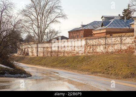 Eine nasse Straße geht hinunter, eine alte Ziegelmauer und Dächer von Häusern auf dem Hügel dahinter. Frühling, Schnee schmilzt, trockenes Gras rundherum, kleine Schneeverwehungen und Pfützen. Tag, bewölktes Wetter, weiches warmes Licht. Stockfoto