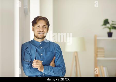 Porträt eines glücklichen jungen Geschäftsmannes, der sich an der Wand lehnt, die Kamera anschaut und lächelt Stockfoto