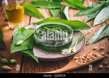 Hausgemachtes Bärlauch-Pesto in einer Glasschüssel auf Holzboden, verziert mit Blättern, Parmesankäse und Olivenöl Stockfoto