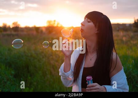 Hübsche junge Frau, die Seifenblasen im Sommer grünes Feld bei Sonnenuntergang. Glückliche friedliche Zeit. Millennial Generation. Stockfoto