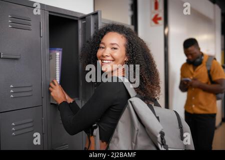 Eine junge Studentin mit gemischter Rasse lächelt und nimmt ihre Bücher auf dem Campus aus dem Schließfach Stockfoto