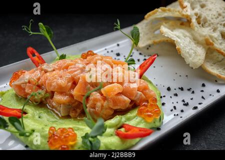 Lachs-Tatar mit rotem Avocado-Püree, Kaviar und Croutons. Auf einem Teller, auf einem dunklen Hintergrund Stockfoto
