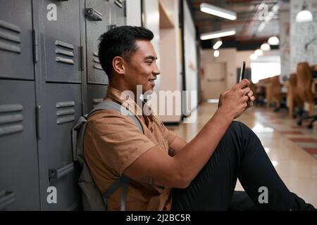 Ein junger Student sitzt auf dem Campus und schreibt seinen Freunden SMS auf seinem Handy Stockfoto