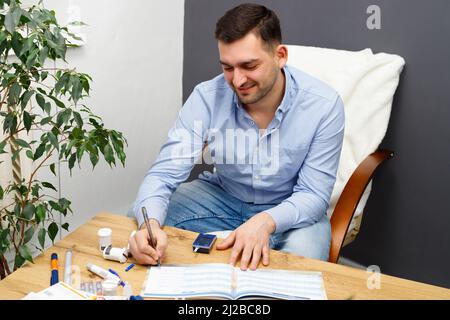 Glücklicher Mann, der zu Hause auf dem Stuhl am Tisch sitzt und Blutzucker in das Tagebuch der Selbstkontrolle schreibt. Stockfoto