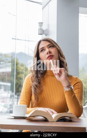 Nachdenkliche junge lateinische Frau mit Make-up, mit Bleistift sitzend, offenes Buch und eine Tasse Kaffee auf dem Tisch, junger Student Lifestyle und Schönheit Stockfoto