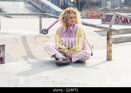 Hübsche junge blonde lockige haarige Frau auf dem grauen Boden sitzen und wegschauen, lächelnd, Beine gekreuzt, ein Roller auf dem Hintergrund Zukunftsperspektiven Konzept voller Schuss Kopie Raum. Hochwertige Fotos Stockfoto