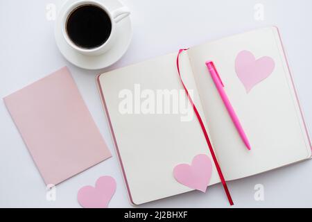 Draufsicht auf Notizbuch mit Papierherzen und einer Tasse Kaffee in der Nähe des Umschlags auf Weiß Stockfoto