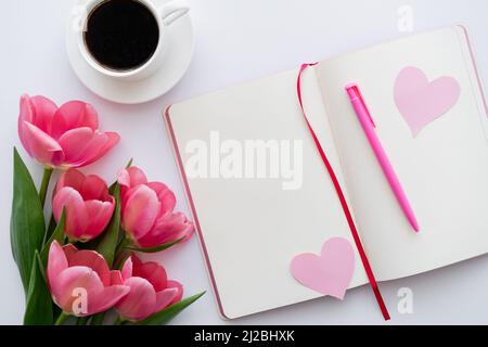 Draufsicht auf Tulpen in der Nähe des Notizbuchs mit Papierherzen und einer Tasse Kaffee auf Weiß Stockfoto