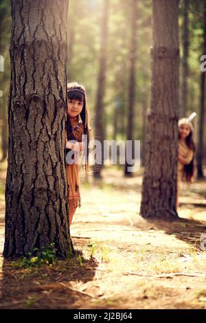 Die Natur ist ein Teil unseres Erbes. Portrait von zwei kleinen Mädchen, die sich hinter Bäumen verstecken, während sie im Wald Dressup spielen. Stockfoto