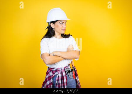 Die junge kaukasische Frau trägt Hardhut und Bauerkleidung auf isoliertem gelben Hintergrund und denkt müde und langweilen mit gekreuzten Armen Stockfoto