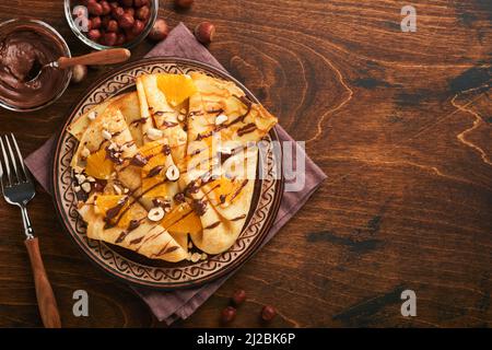 Crepes-Suzette mit Orangen und Schokolade. Dünne Crepes mit Schokoladenaufstrich, Haselnüssen und Orangenscheiben Obst auf dem Teller zum Frühstück auf altem dunklen Woo Stockfoto