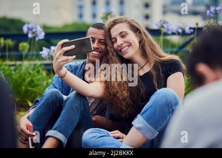 Erinnerungen mit meinen Bindungen machen. Aufnahme von zwei jungen Freunden, die zusammen ein Selbstporträt machen, während sie tagsüber in einem Park draußen sitzen. Stockfoto