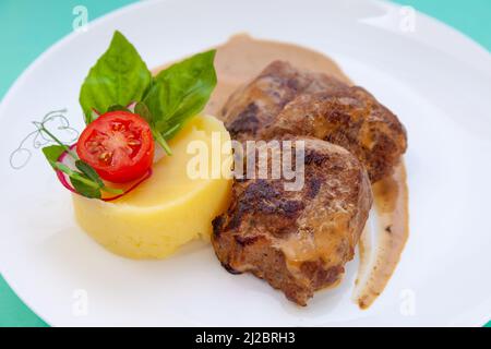 Fleischbällchen mit Kartoffelpüree auf einem weißen Teller. Fleisch in saurer Rahmsoße. Tomaten und Basilikum. Essen im Restaurant. Mittagspause. Medaillons. Stockfoto
