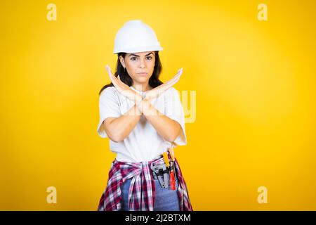 Junge kaukasische Frau trägt Hardhut und Bauerkleidung über isoliertem gelben Hintergrund Ablehnung Ausdruck Kreuzung Arme tun negative Zeichen, Angr Stockfoto