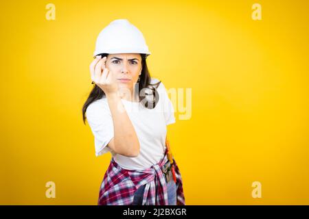 Junge kaukasische Frau mit Hardhut und Bauerkleidung auf isoliertem gelbem Hintergrund, die Capice oder Geldgeste macht und Ihnen sagt, dass Sie Ihre Schulden bezahlen sollen Stockfoto