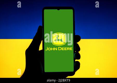 In dieser Abbildung ist ein John Deere-Logo auf einem Smartphone mit einer ukrainischen Flagge im Hintergrund zu sehen. Stockfoto