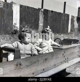 1961, draußen auf einem Spielplatz, zwei kleine Mädchen sitzen in einem hölzernen gerahmten Schaukelpferd, England, Großbritannien. Der erste Spielplatz der Welt stammt aus der Mitte des Jahres 1800s, wo ein Spielbereich für Kinder geschaffen wurde, um mit Holzblöcken und Spielzeug zu spielen. Der Spielplatz, wie wir ihn heute kannten, entwickelte sich als eine Möglichkeit, Kinder abseits der Straßen zu ermutigen, und ab dem Jahr 1930s wurden aus Holz und Metall gefertigte Strukturen wie Rutschen, Schaukeln, Klettergerüste und Ausritte als Möglichkeit für Kinder geschaffen, sich draußen zu amüsieren. Mit der Einführung neuer Gesundheits- und Sicherheitsvorschriften wurden solche Strukturen aus dem Jahr 1980s entfernt. Stockfoto