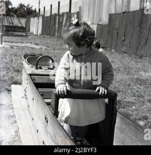 1961, draußen auf einem Spielplatz, ein kleines Mädchen, das in einem hölzernen Schaukelpferd steht, England, Großbritannien. Ein Hexenhut ist ebenfalls zu sehen. Der weltweit erste Spielplatz stammt aus der Mitte des Jahres 1800s, ein Bereich, in dem Kinder mit Holzblöcken und Spielzeug spielen können. Der Spielplatz, wie wir ihn heute kannten, entwickelte sich als eine Möglichkeit, Kinder abseits der Straßen zu ermutigen, und ab dem Jahr 1930s waren Holz- und Metallkonstruktionen wie Rutschen, Schaukeln, Klettergerüste und Ausritte für die Kinder nicht mehr geeignet, sich in einer sicheren Umgebung zu amüsieren. Die Gesundheits- und Sicherheitsvorschriften sahen Ende 1980s die Entfernung solcher Strukturen vor. Stockfoto