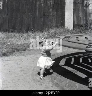 1961, historisch, draußen auf einem öffentlichen Spielplatz, ein kleines Mädchen, das sich am Rand eines hölzernen Kreisels oder eines Karussels festhält, England, Großbritannien. Es wird vermutet, dass der erste Spielplatz der Welt auf die Mitte 1800s zurückgeht, wo ein Bereich für Kinder geschaffen wurde, um mit Holzblöcken und Spielzeug zu spielen. Der Spielplatz entwickelte sich dezent als eine Möglichkeit, Kinder abseits der Straßen zu ermutigen und ab dem 1930s aus Holz und Metall gefertigte Rutschen, Schaukeln, Klettergerüste und Ausritte boten Kindern nach Viel Spaß. einen Auslauf Mit dem Anstieg der Gesundheits- und Sicherheitsvorschriften wurden solche Strukturen aus dem Jahr 1980s entfernt Stockfoto