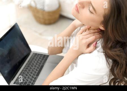 Müde junge Frau mit Nackenschmerzen, die im Büro zu Hause in einem Laptop arbeitet. Müde junge Frau berührt ihren steifen Hals, fühlt Gelenkschmerzen, Rückenschmerzen, massiert Stockfoto
