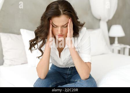 Unglückliche Frau, die ihren Kopf berührt, zu Hause auf dem Bett sitzt und Kopfschmerzen hat. Junge frustrierte Frau denkt über Probleme nach, fühlt sich einsam und traurig. Stockfoto