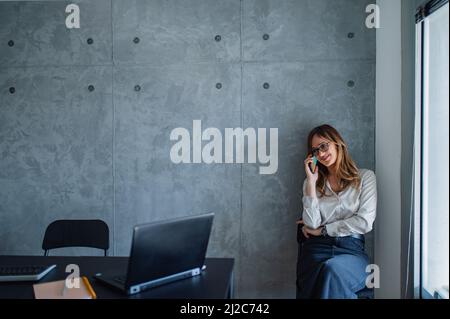 Eine Frau mittleren Alters mit Denim-Rock und weißem Hemd sitzt auf einem Stuhl in der Ecke des Arbeitsplatzes und macht Pause, spricht am Telefon und entspannt sich. Stockfoto
