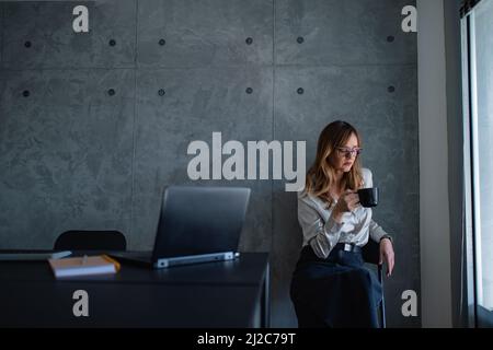 Eine kaukasische Geschäftsfrau mit Brille sitzt auf einem Stuhl in der Ecke des modernen grauen Büros des Unternehmens und schaut auf die schwarze Tasse Kaffee oder Tee. Stockfoto