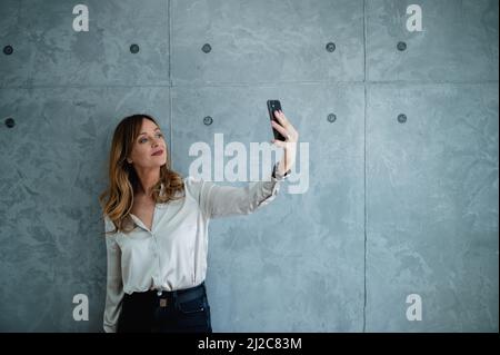 Eine Erwachsene kaukasische Frau mit grünen Augen macht Selfies mit ihrem Smartphone. Sie steht in ihrem Büro vor einer grauen Wand. Stockfoto