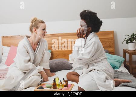 Zwei junge Frauen in Masken sitzen neben einem Set für Spa-Behandlungen. Die Frauen in Bademänteln sitzen auf dem Bett und schauen auf die Kamera Stockfoto