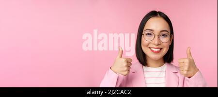 Super, Glückwunsch. Gesicht einer aufgeregten asiatischen Geschäftsfrau in einer Brille, lächelnd zufrieden, mit Daumen nach oben, stehend über rosa Hintergrund Stockfoto