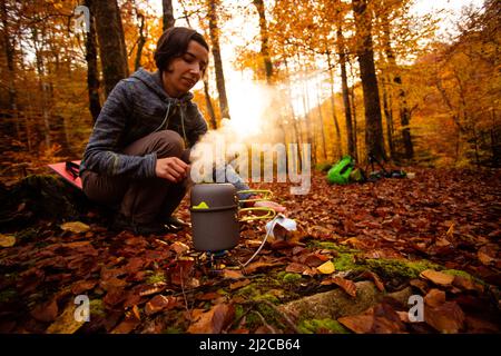 Frau verwendet tragbare Gasheizung und Pfanne zum Kochen im Freien während des Campens. Zusammenklappbare Flaschen, um Platz in Ihrem Rucksack zu sparen. Stockfoto