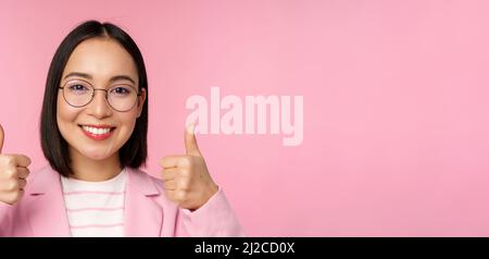 Super, Glückwunsch. Gesicht einer aufgeregten asiatischen Geschäftsfrau in einer Brille, lächelnd zufrieden, mit Daumen nach oben, stehend über rosa Hintergrund Stockfoto