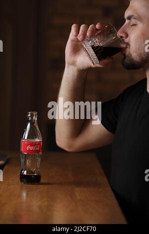 RIVNE, UKRAINE JUNI 26 2020: Junger bärtiger Mann trinkt im Café Coca Cola aus Glas. Flasche mit Cola steht auf einem Holztisch. Stockfoto